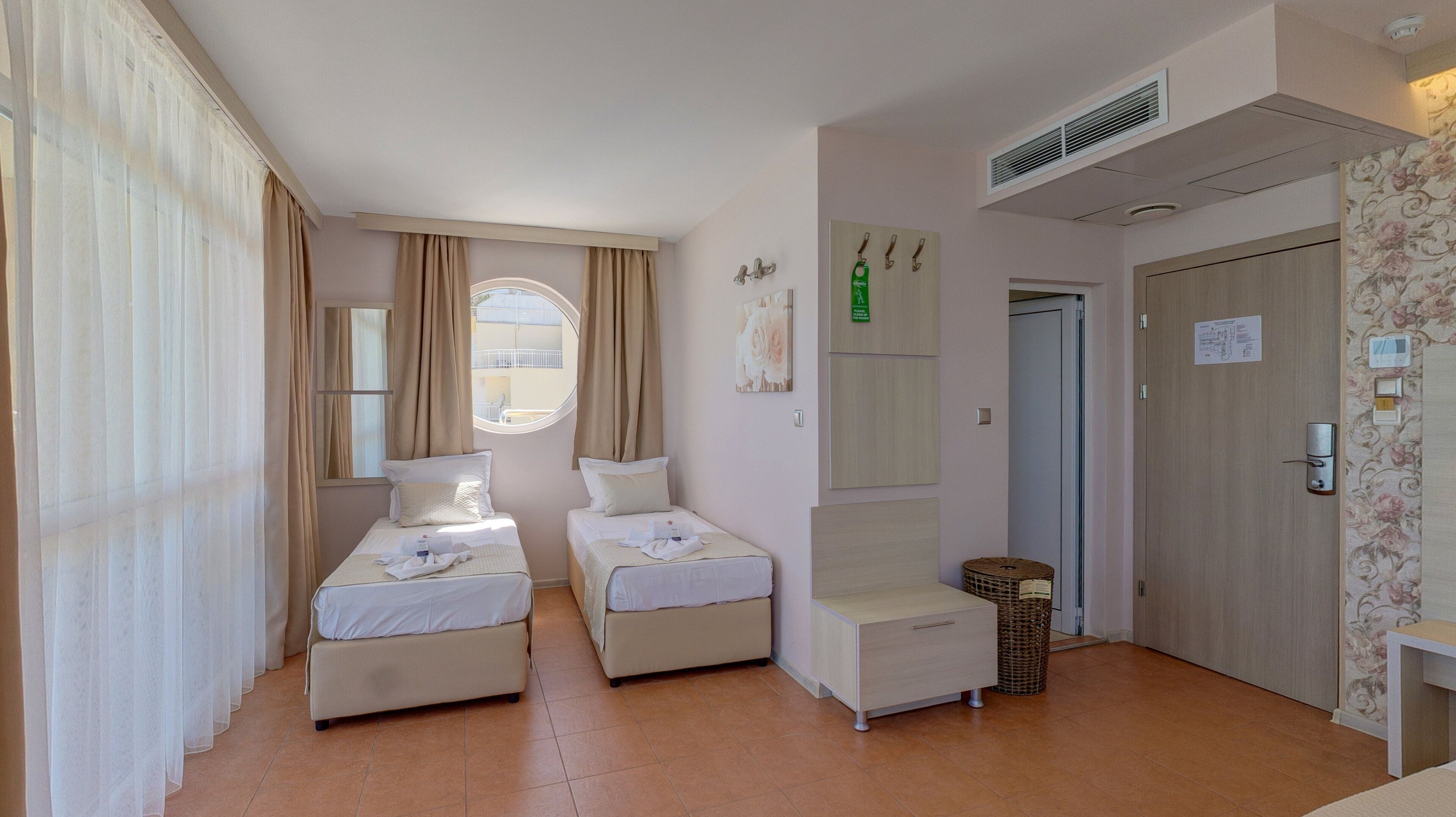 Une chambre d’hôtel avec deux lits simples, une petite table, une chaise, une fenêtre avec des rideaux, une porte avec une pancarte et une bouche d’aération fixée au mur.