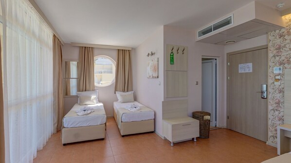 A hotel room with two single beds, a small table, a chair, a window with curtains, a door with a sign, and a wall-mounted air vent. - Aphrodite Beach Hotel (Nessebar)