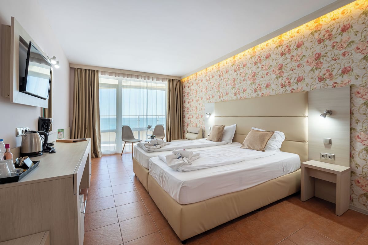 A hotel room with two beds, a desk with a kettle, a chair, and a view of the outdoors.