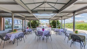 Un espace extérieur aménagé en terrasse, doté de plusieurs tables et chaises, abrité par un toit et entouré de verdure.