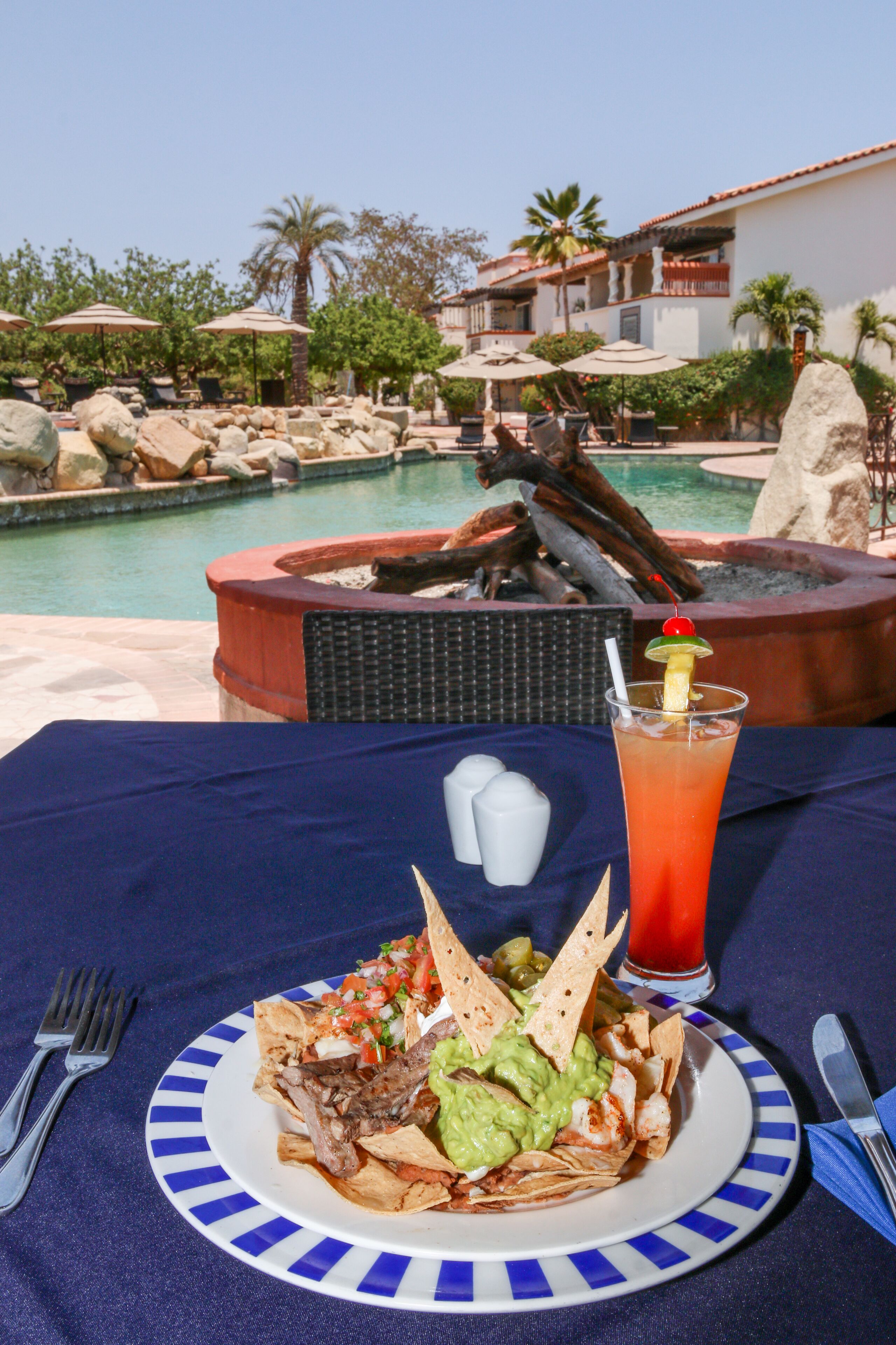 A dining table with a plate of nachos, a drink, and a view of a pool and outdoor seating area.