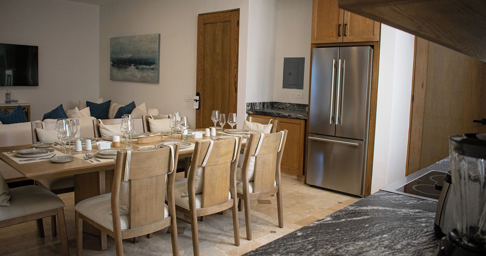 Un comedor con una mesa de madera dispuesta para seis personas, rodeada por seis sillas. Se observa un refrigerador de acero inoxidable al fondo.