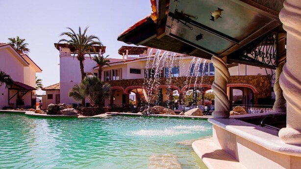 Un área de piscina con una fuente, palmeras y edificios con paseos arqueados.