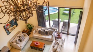 Amplia sala de estar con un gran candelabro, un conjunto de sofá blanco, una mesa de centro y vistas a un patio exterior con piscina.