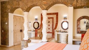 Una habitación de hotel con un arco de piedra, una cama grande con una colcha naranja, una puerta de madera y un baño con espejo y lavamanos.