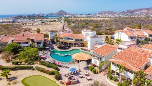 Un resort con piscina, varias edificaciones con techos de tejas rojas y vistas al océano y montañas al fondo.