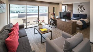 Una habitación de hotel moderna con sofá, escritorio con laptop, cama y ventana grande con vista a la ciudad.