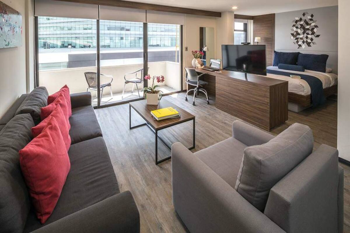 Una habitación de hotel moderna con sofá, escritorio con laptop, cama y ventana grande con vista a la ciudad.