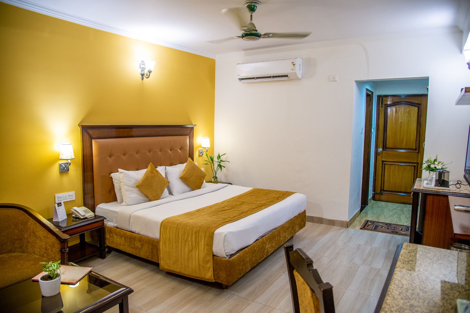 Habitación de hotel con una cama grande, un escritorio, una silla, una mesita de noche, una lámpara, aire acondicionado y una puerta.