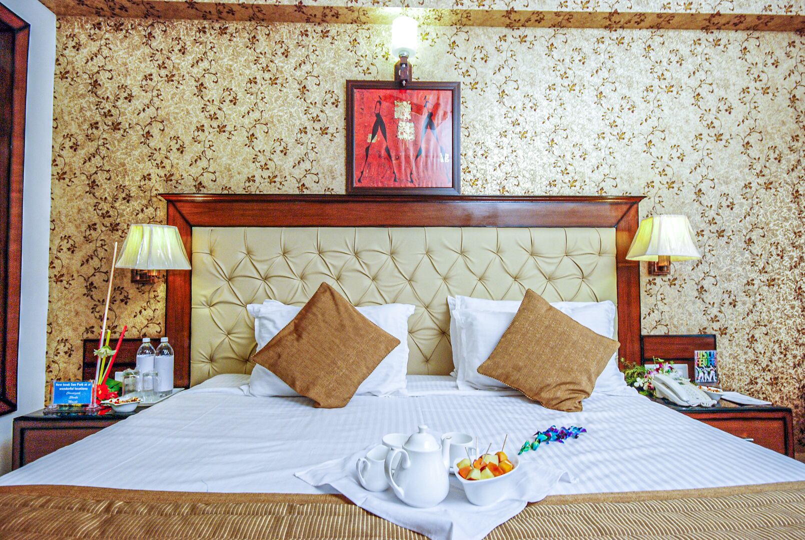 Ein Hotelzimmer mit einer gebügelten Kopfteil, blumigem Wandpapier, zwei Nachttischlampen, einem gerahmten Kunstwerk über dem Bett und einem Frühstücksbrettchen auf dem Bett.
