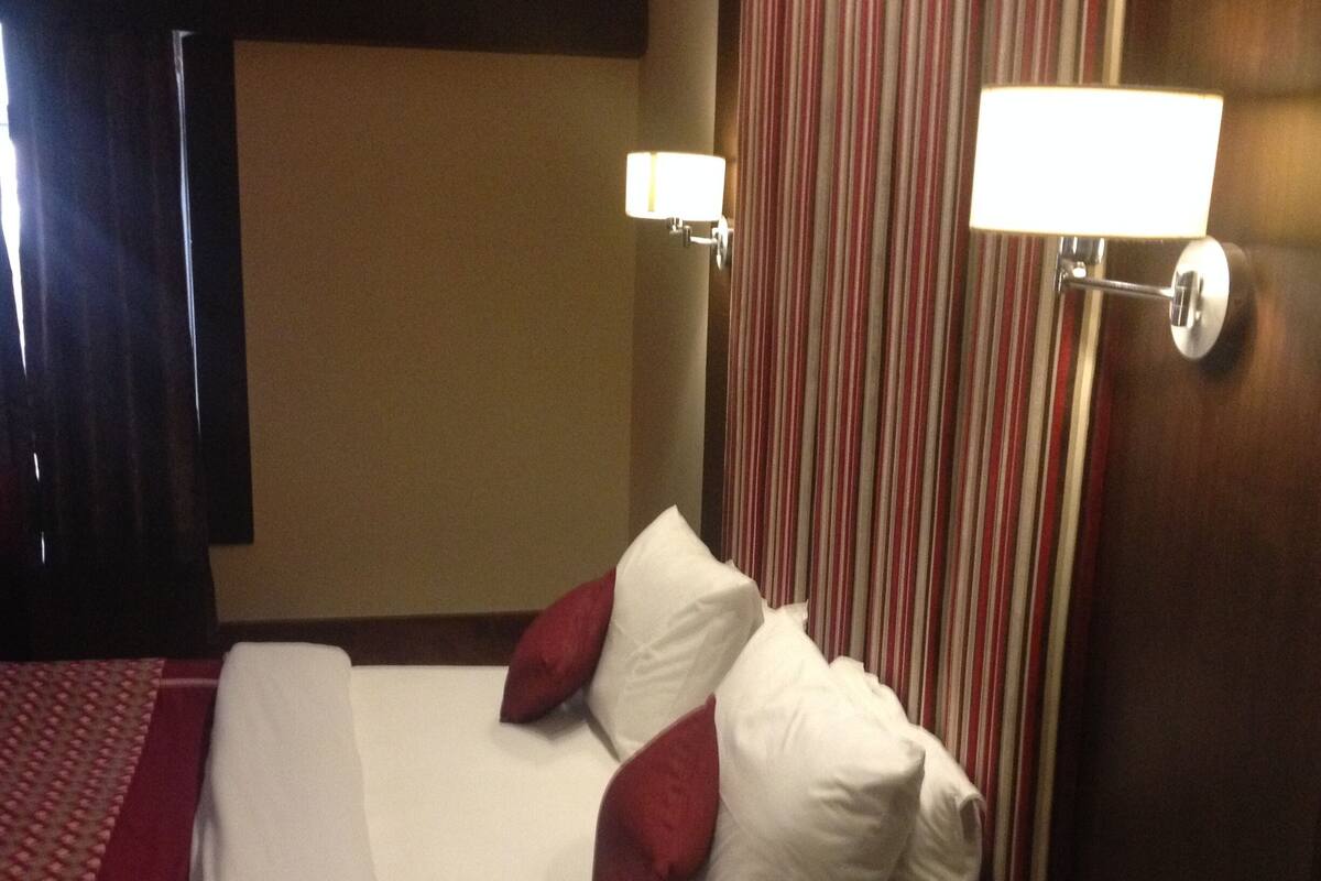 A hotel room with a bed, pillows, a nightstand, a lamp, and a striped curtain.