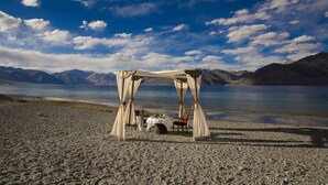 A beach setting with a canopy and a table set for dining, overlooking a calm lake and mountains. - The Grand Dragon Ladakh (Leh)