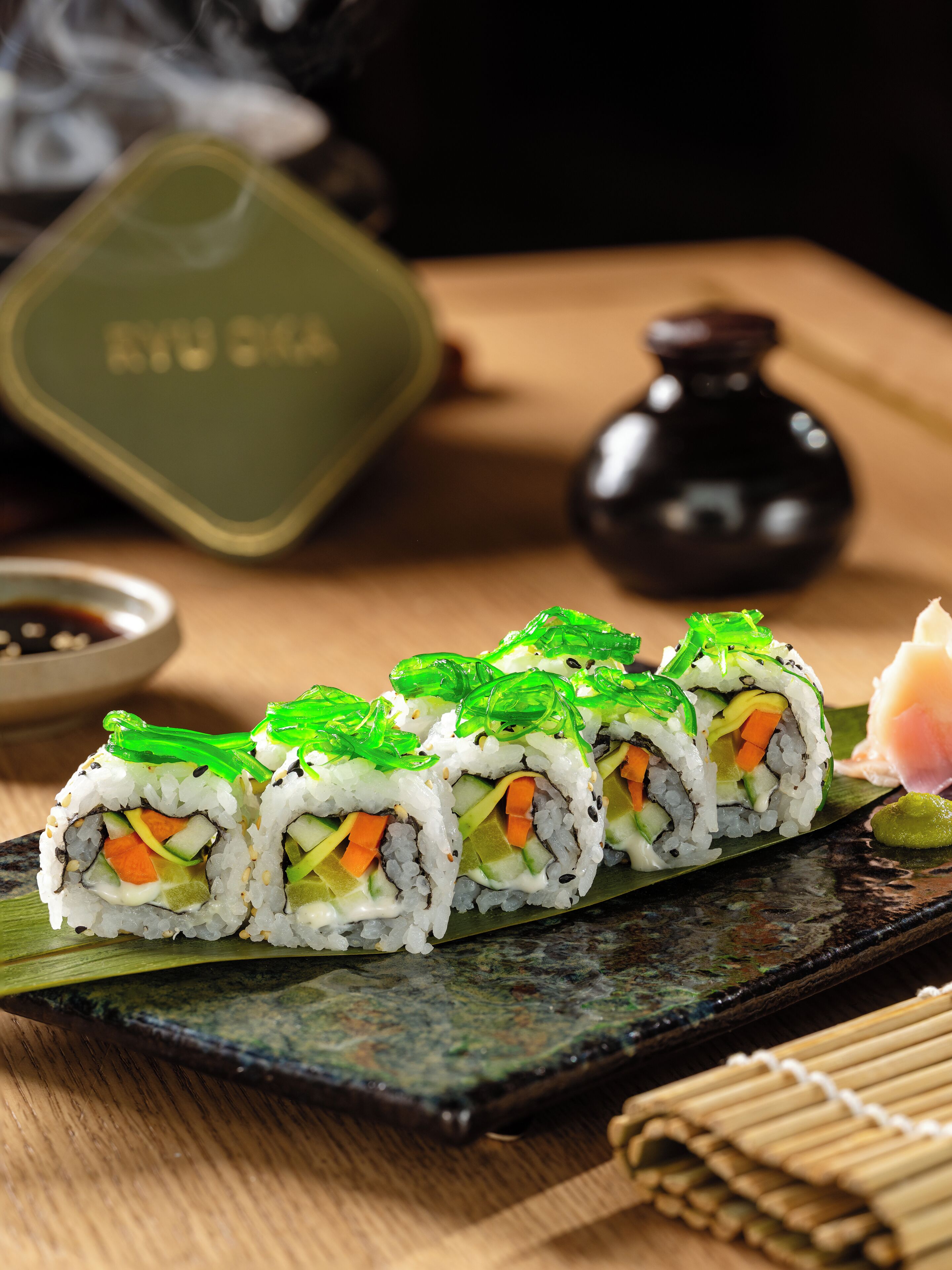 Sushi rolls on a wooden table with a green container and a small black bottle in the background.