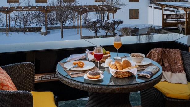 A dining table with food and drinks, a view of snow-covered mountains, and traditional architecture. - The Grand Dragon Ladakh (Leh)