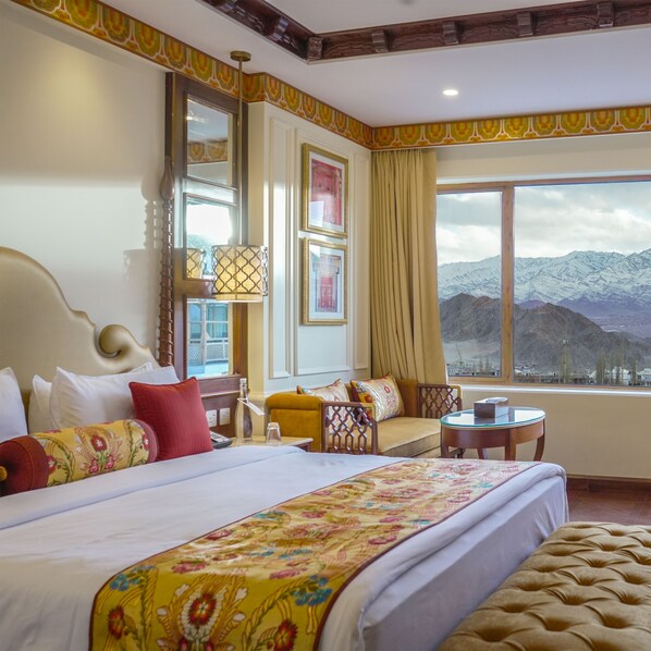 A hotel room with a large bed, a sofa, a small table, and a view of mountains through the window. - The Grand Dragon Ladakh (Leh)