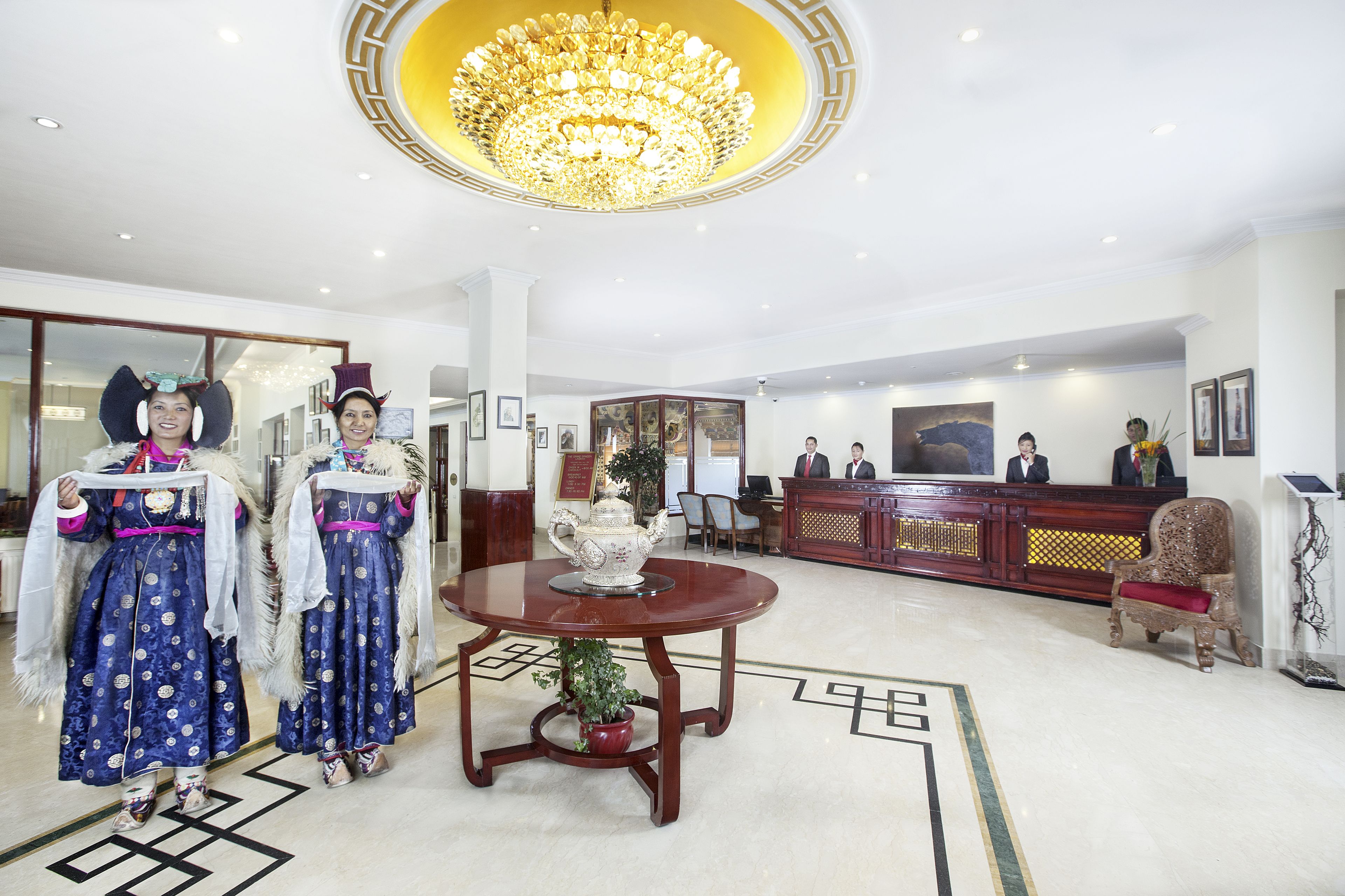 A hotel lobby with a reception desk, a chandelier, and two individuals in traditional attire.