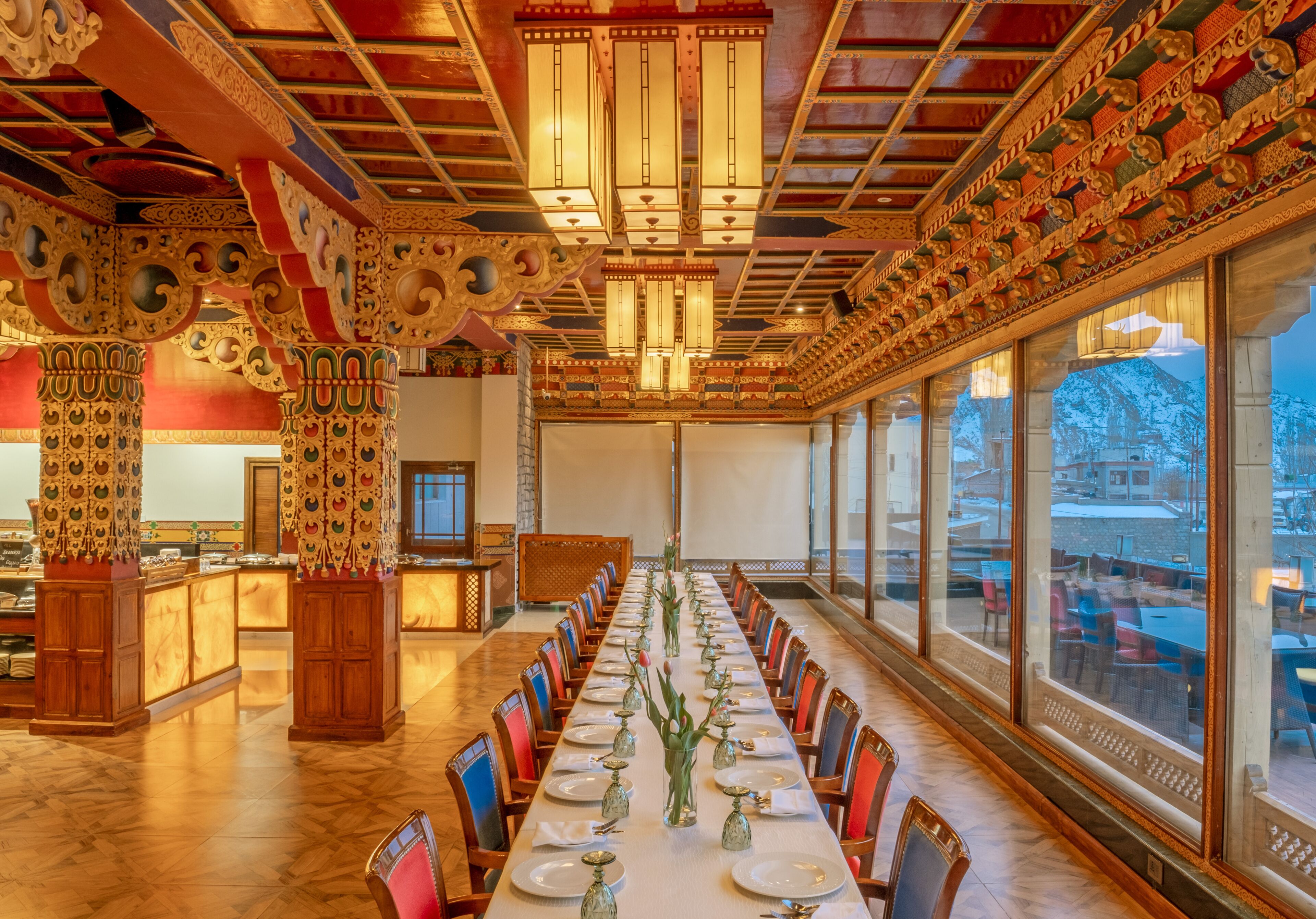 Een lange eettafel met stoelen, een versierd plafond en grote ramen.