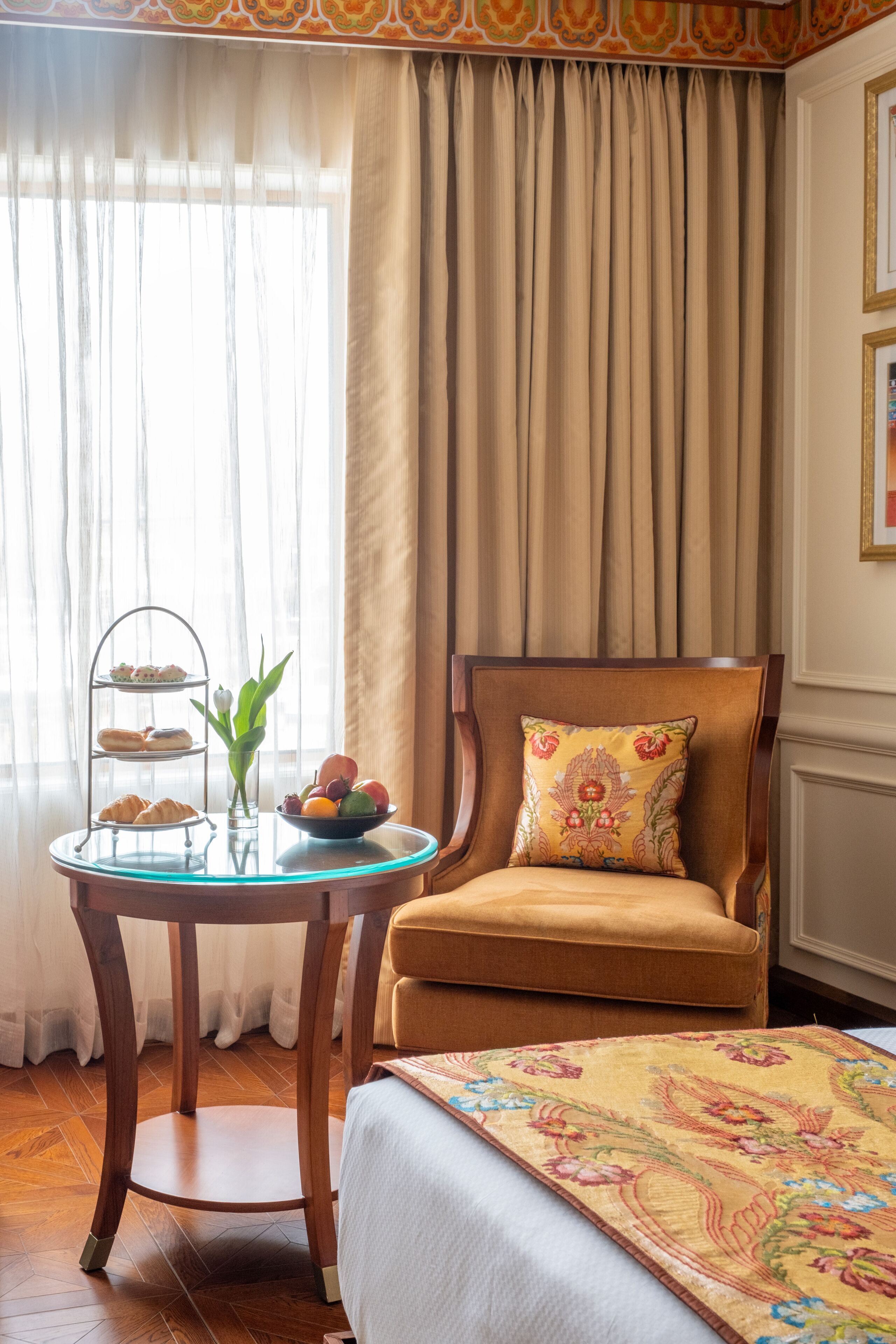 A room with a wooden chair, a small table with a tray of pastries, a bowl of fruit, and a bed with a floral bedspread.
