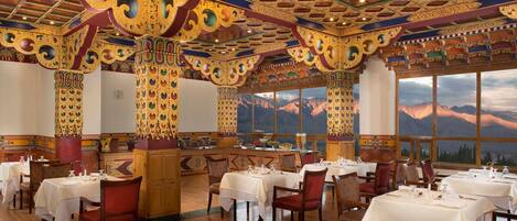 A restaurant with ornate golden columns, wooden tables with white tablecloths, and a mountain view through large windows.