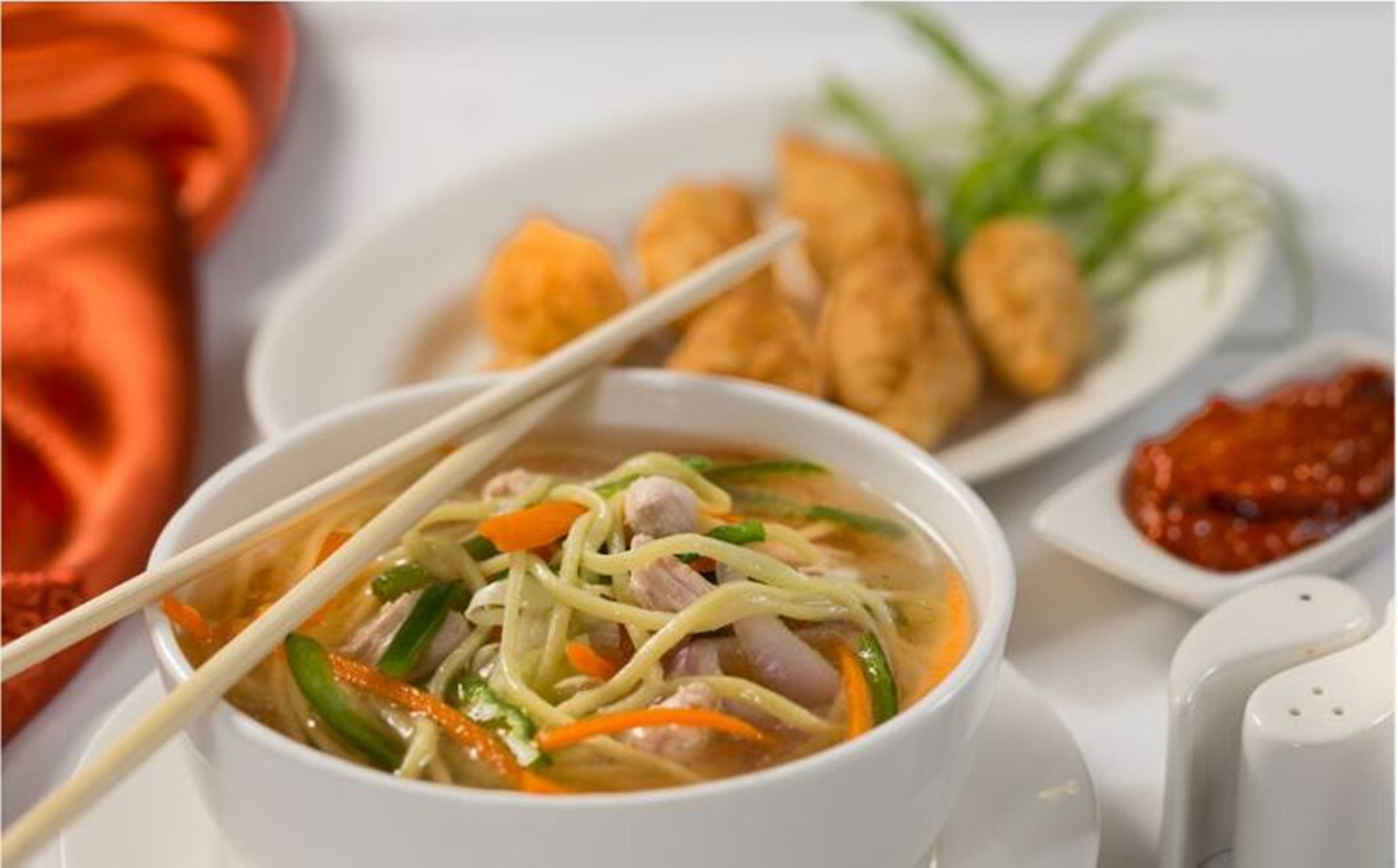Un tazón de sopa con fideos, verduras y carne, acompañado de alimentos fritos y un recipiente pequeño con salsa.