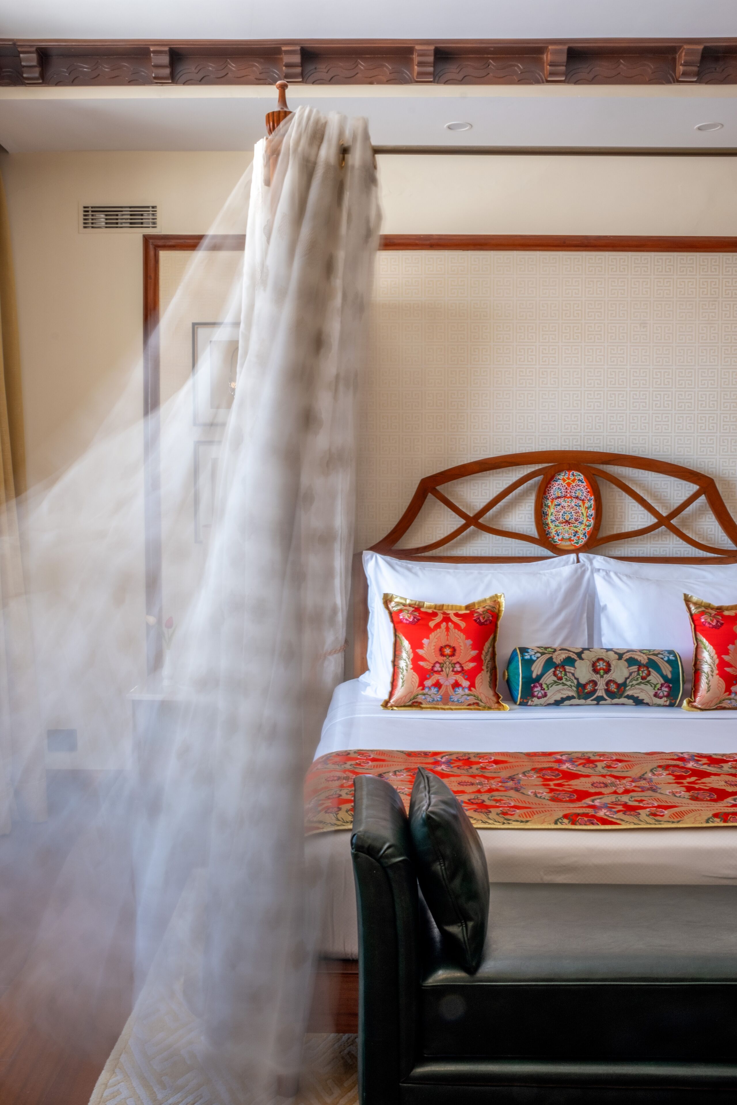 A bed with a patterned headboard, a decorative pillow, and a sheer curtain.