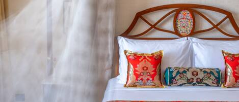 A bed with a patterned headboard, a decorative pillow, and a sheer curtain.