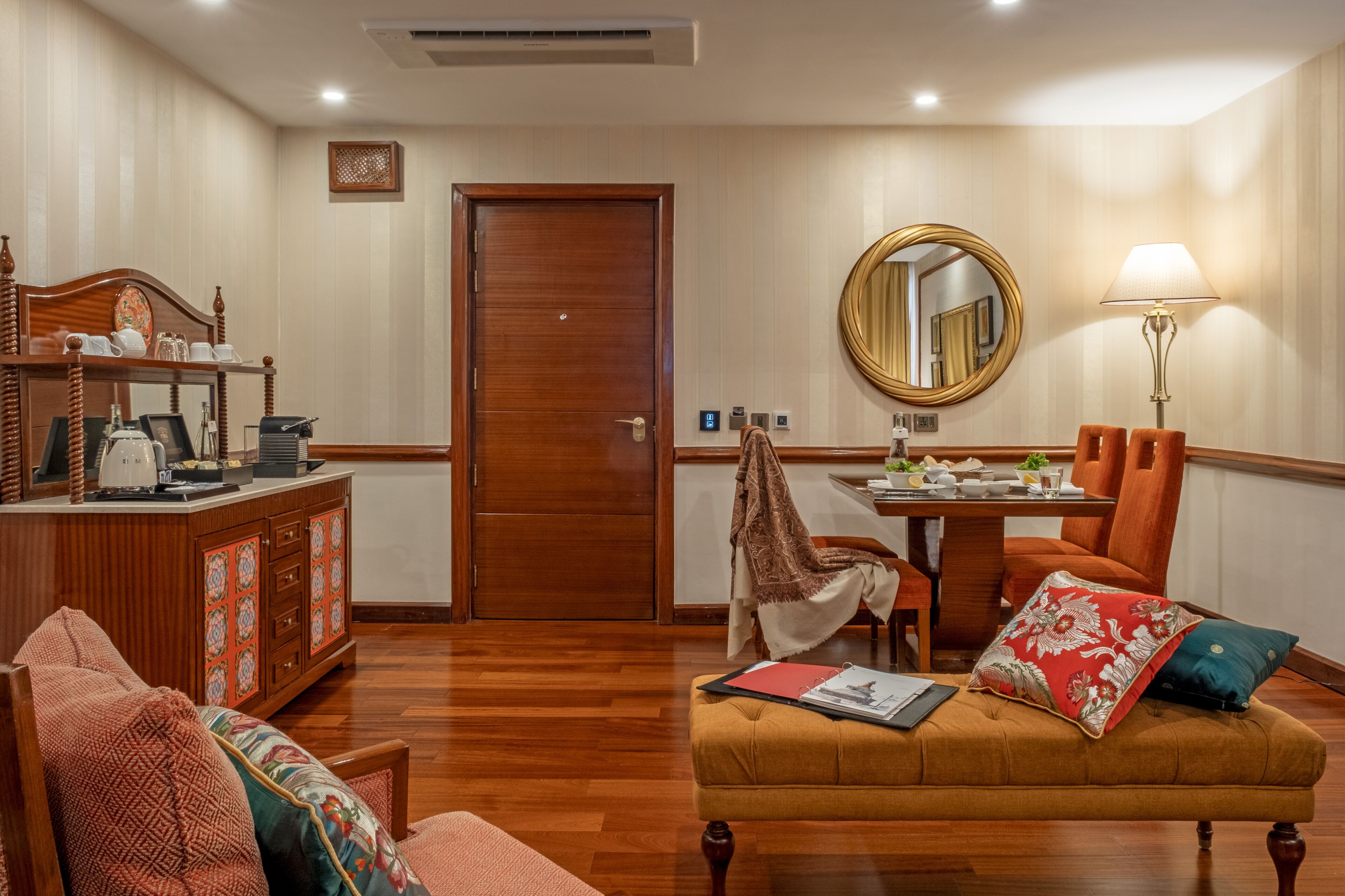 A hotel room with a wooden floor, a dining table with chairs, a sofa, a cabinet, a mirror, and a lamp.