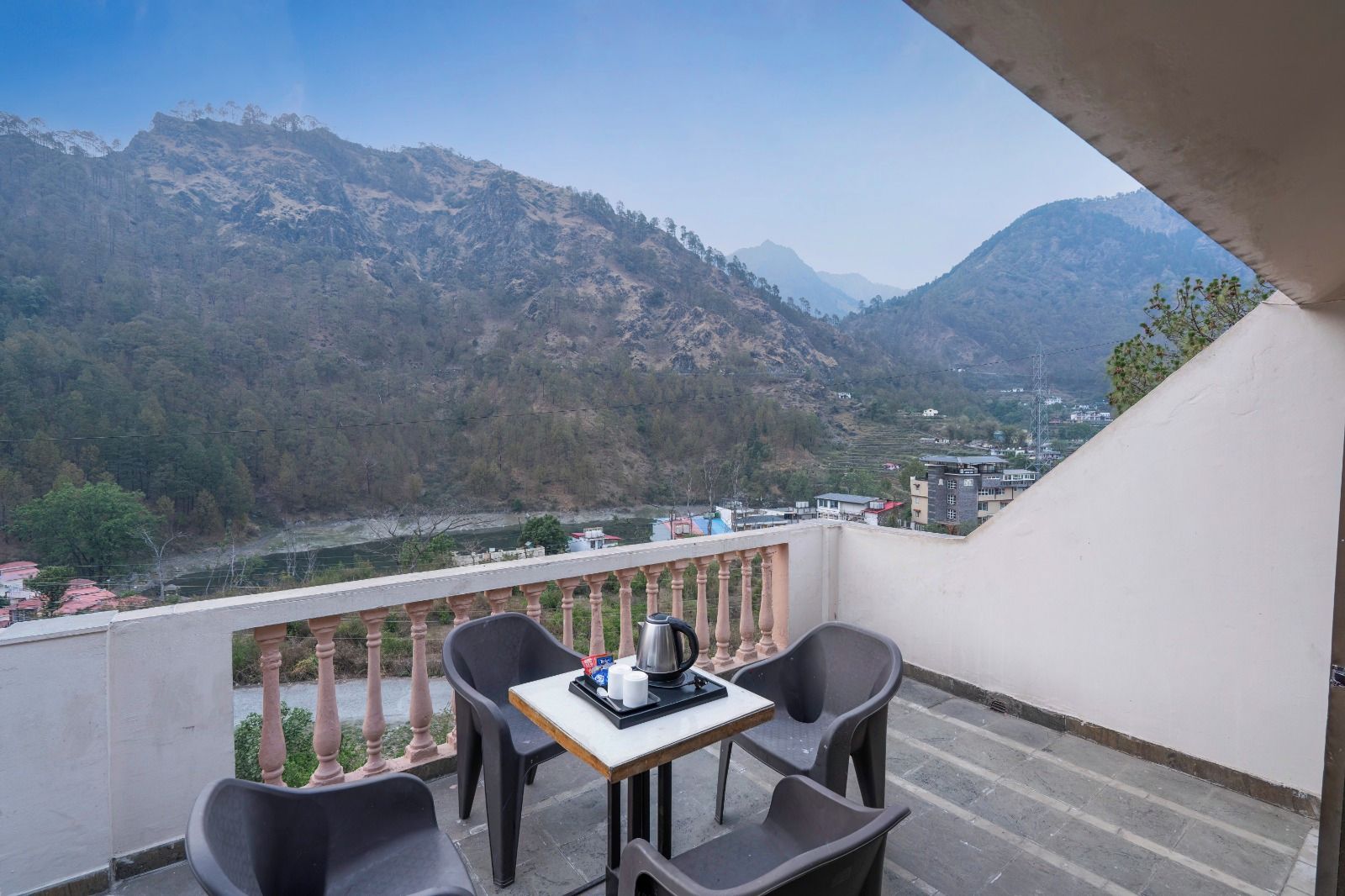 A balcony with a table and chairs, overlooking a mountainous landscape with a river and buildings in the distance.