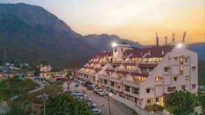 Un hotel de varios pisos con balcones, estacionamiento y montañas al fondo.