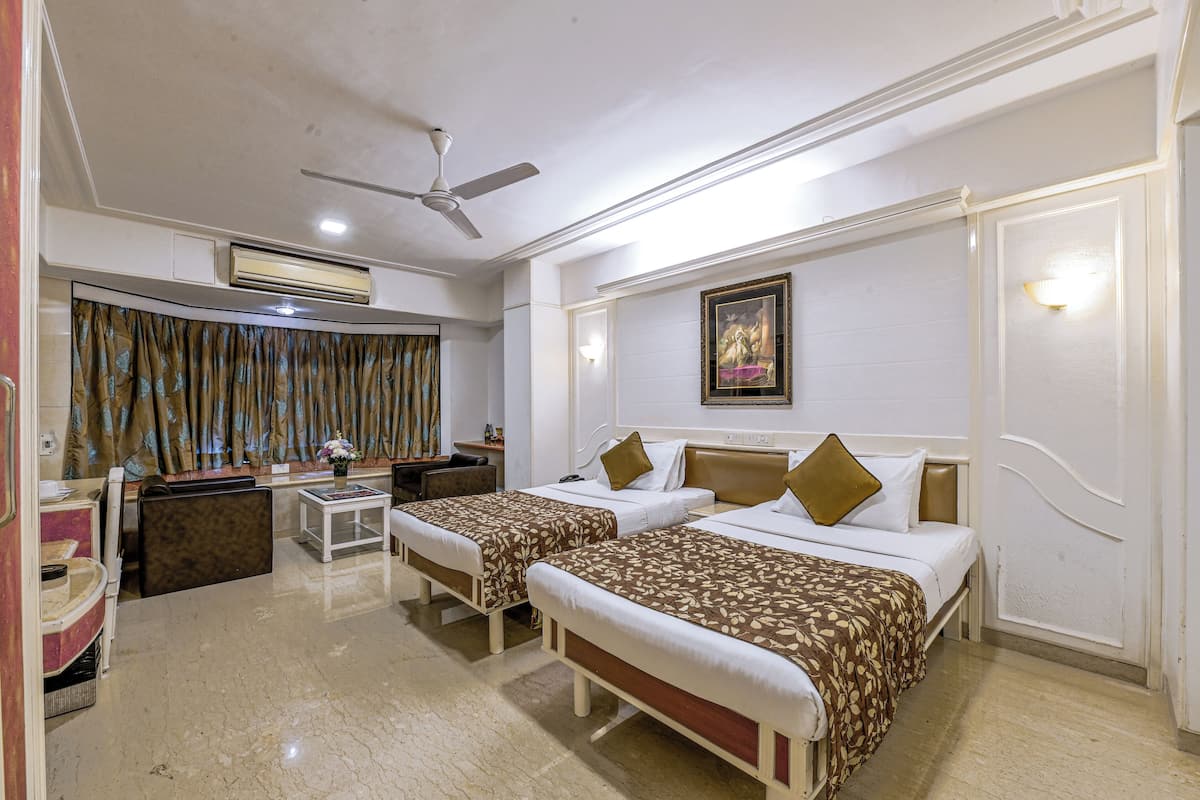 A hotel room with two beds, a ceiling fan, an air conditioning unit, a desk with a chair, a small table with a vase, and a curtain.
