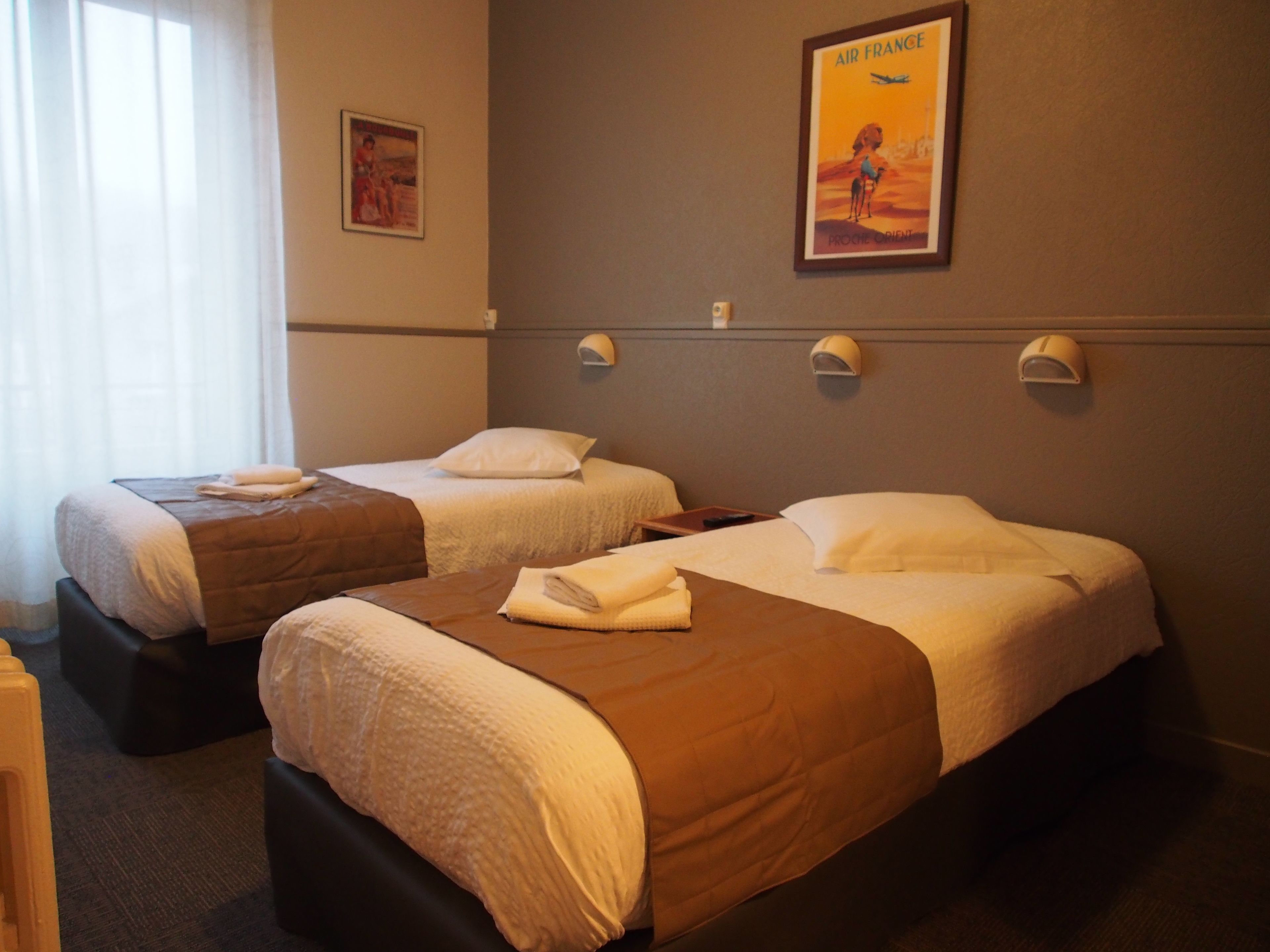 A hotel room with two single beds, each with a pillow and a folded towel. There are wall-mounted lamps on the wall above the beds.