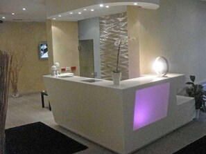 A reception desk with a modern design, a potted plant, and a wall-mounted light fixture. - Trieste Hotel (Rimini)