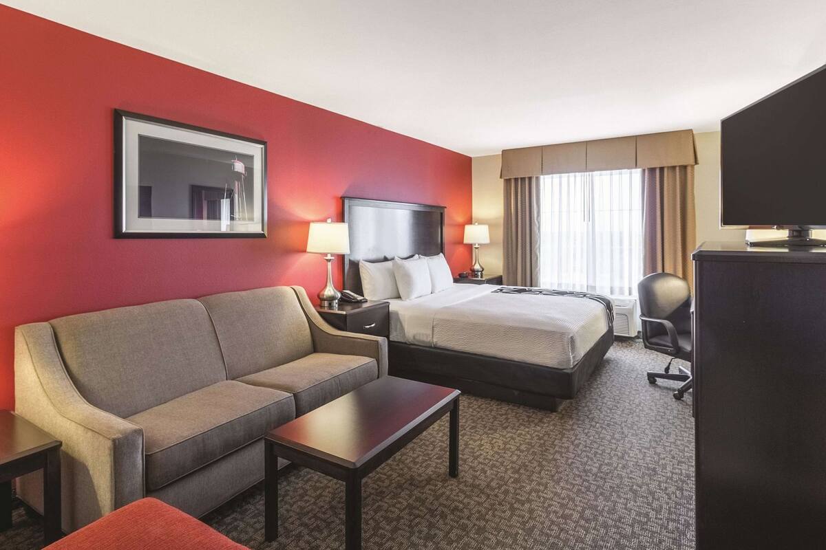 Quarto de hotel com cama, sofá, escrivaninha, televisão e janela com cortinas.
