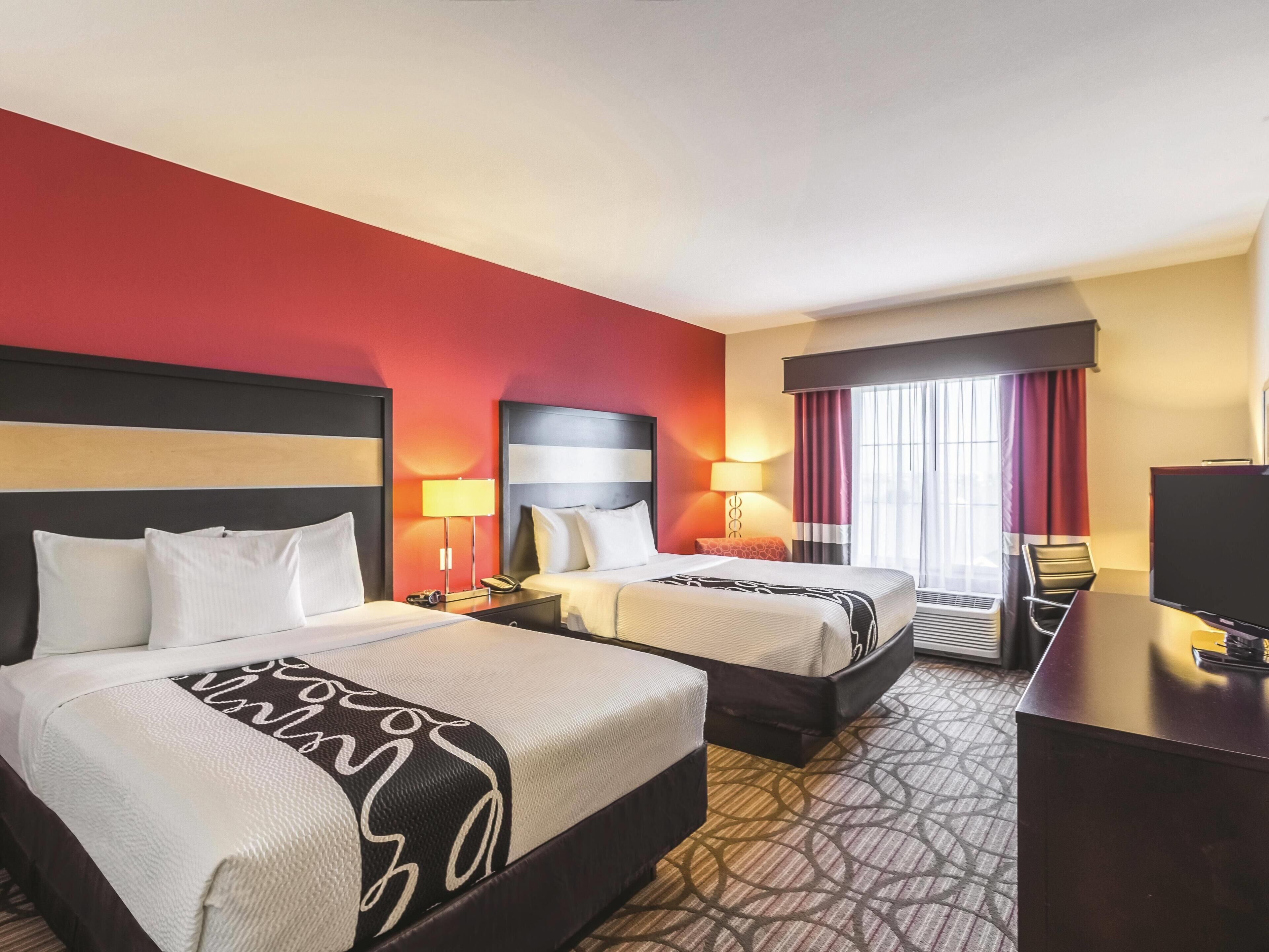 Quarto de hotel com duas camas, uma escrivaninha, televisão e janela com cortinas.