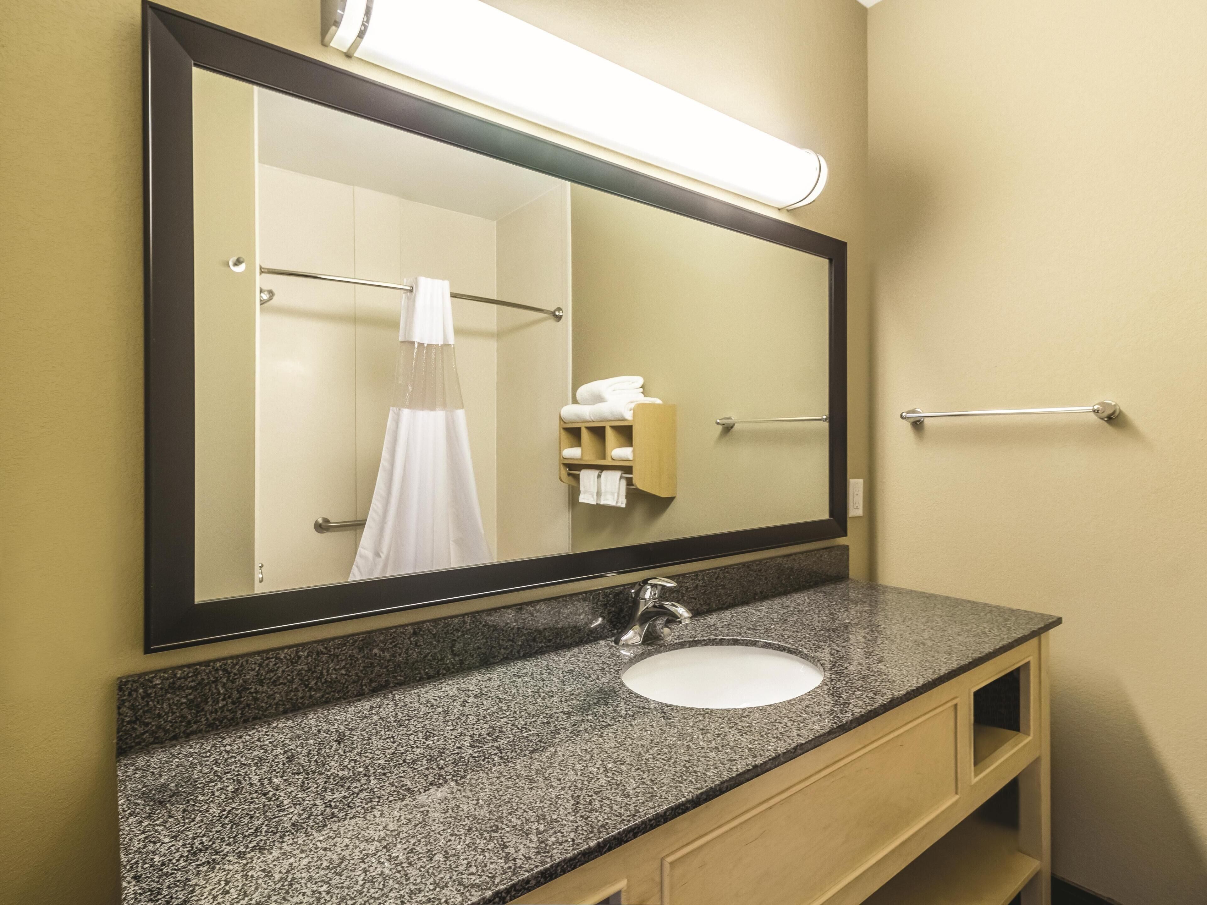 A bathroom with a granite countertop, a sink, a mirror, a towel rack, and a towel dispenser.