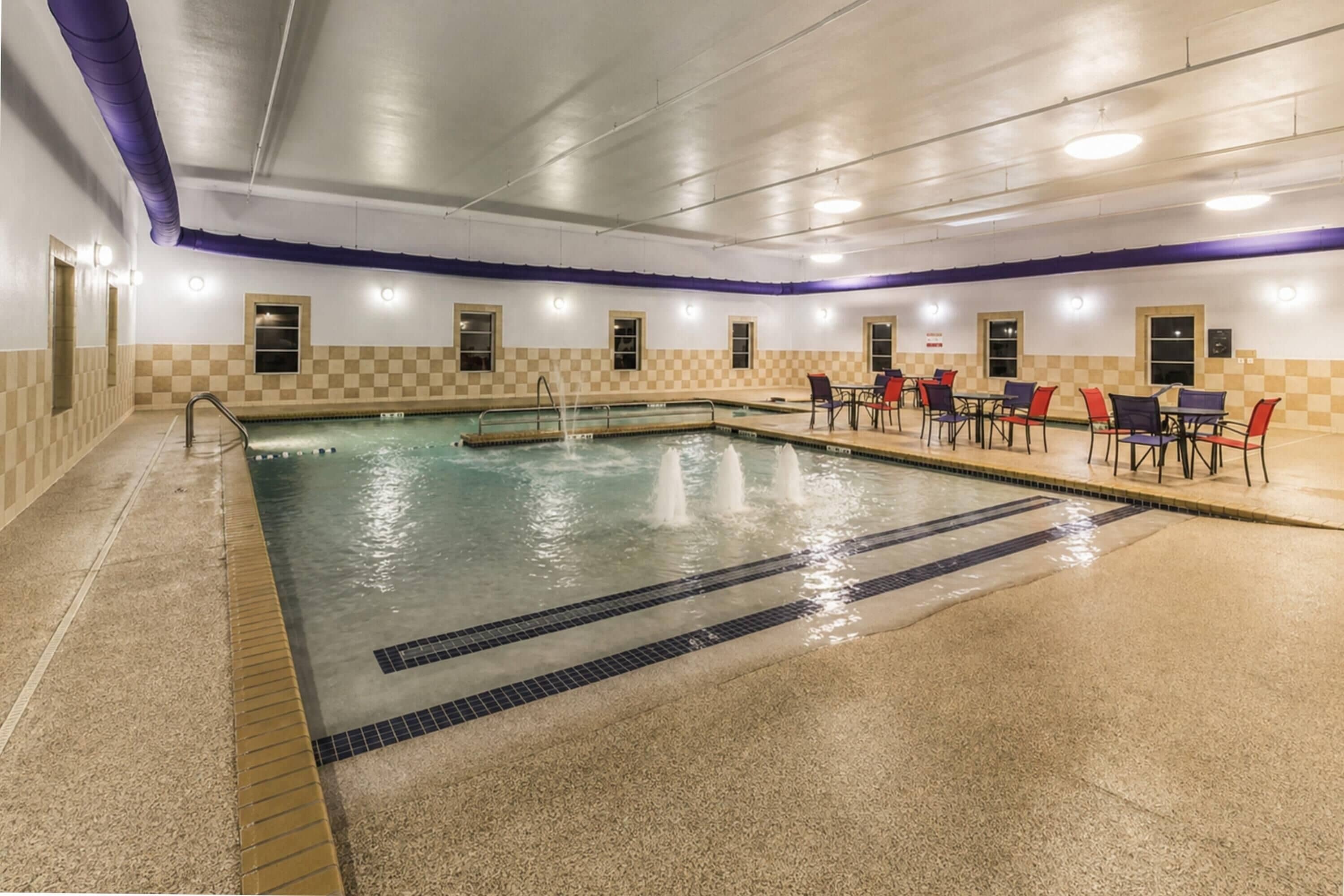 Una piscina interior con diseño de pared a cuadros, una zona de estar con mesas y sillas, y un suelo de baldosas.