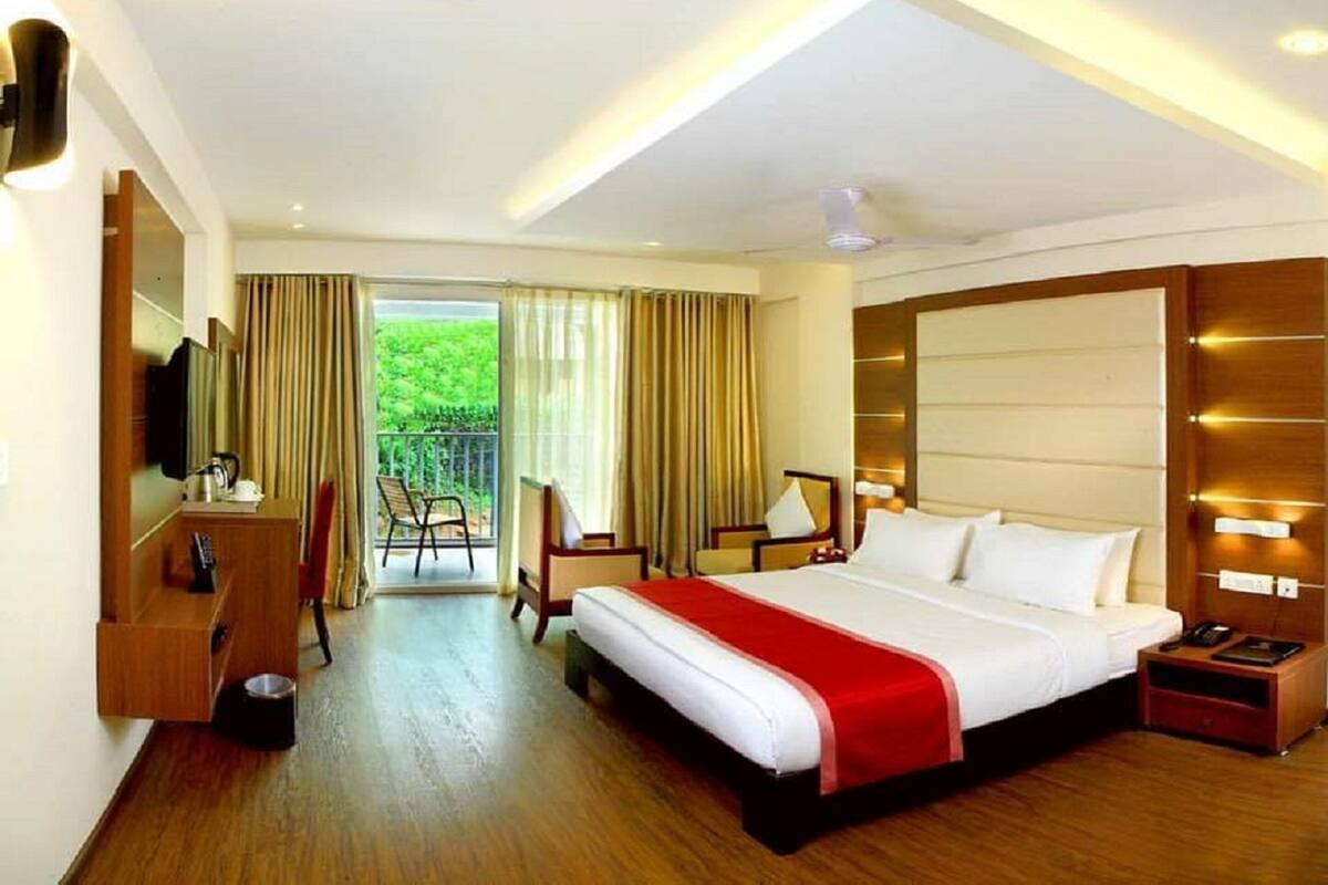 A hotel room with a large bed, a desk, a chair, a TV, and a balcony with curtains.