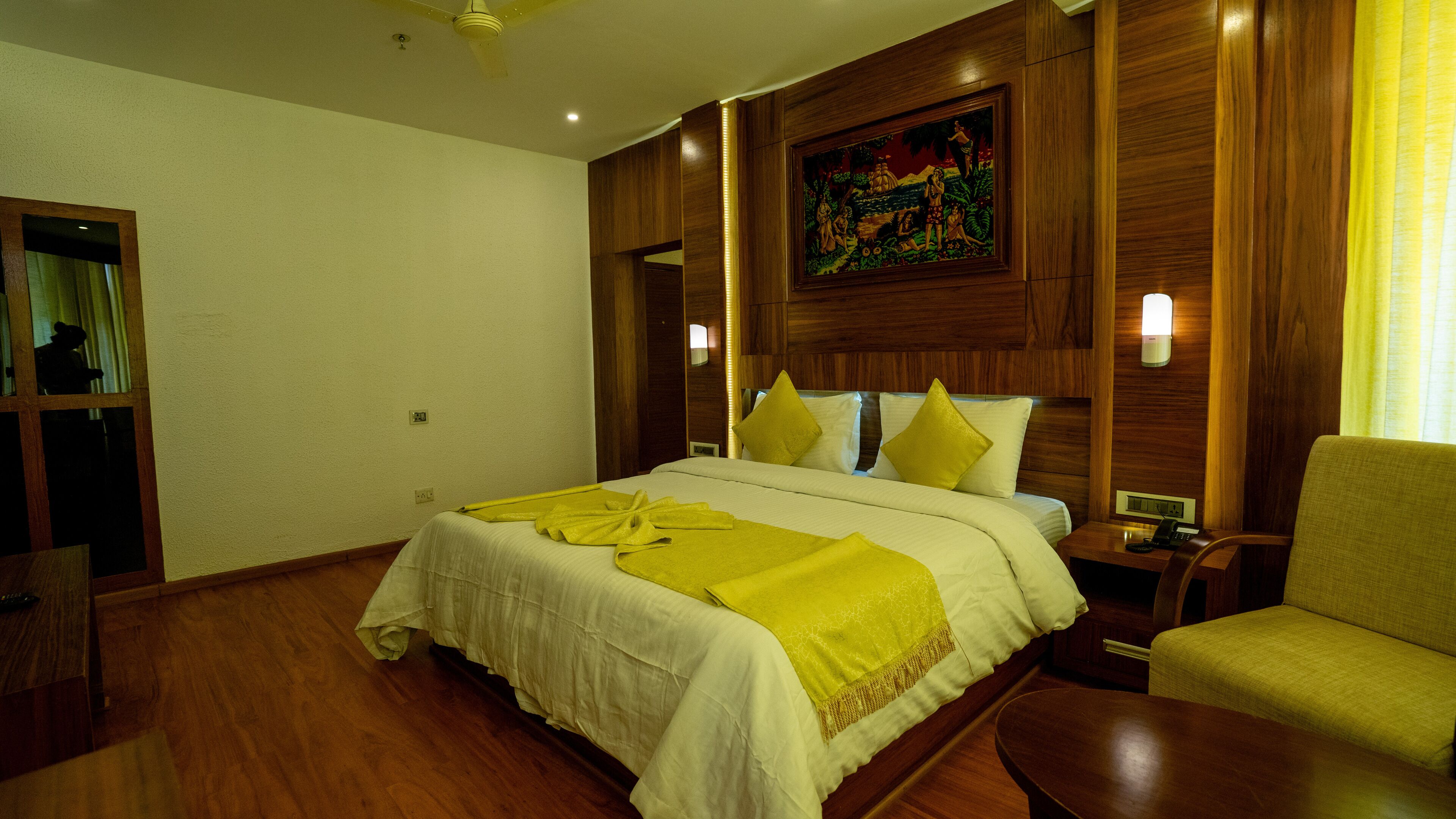 Una habitación de hotel con una cama grande, paneles de madera, un cuadro en la pared y un sillón amarillo.