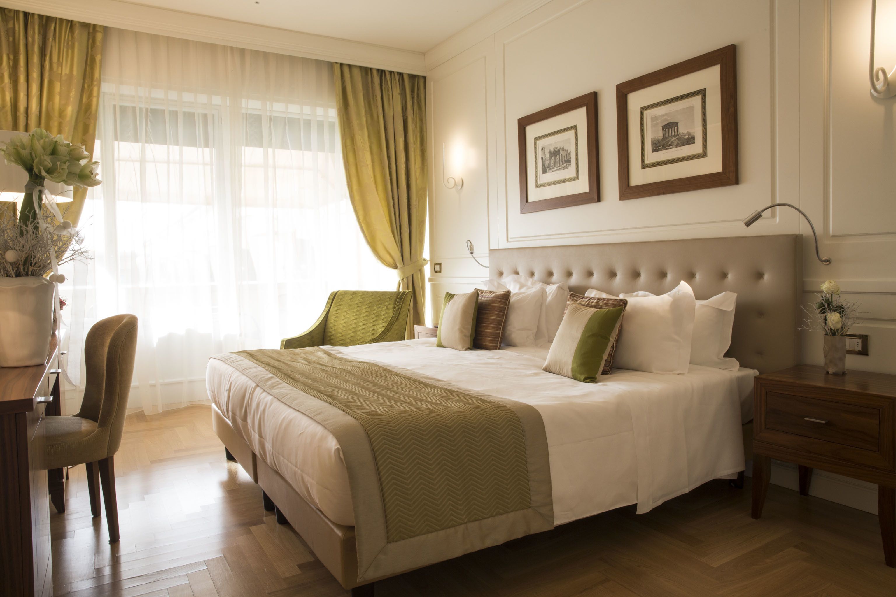 A neatly arranged hotel room with a large bed, a desk, and a chair. There are two framed pictures on the wall and a vase with flowers on the desk.