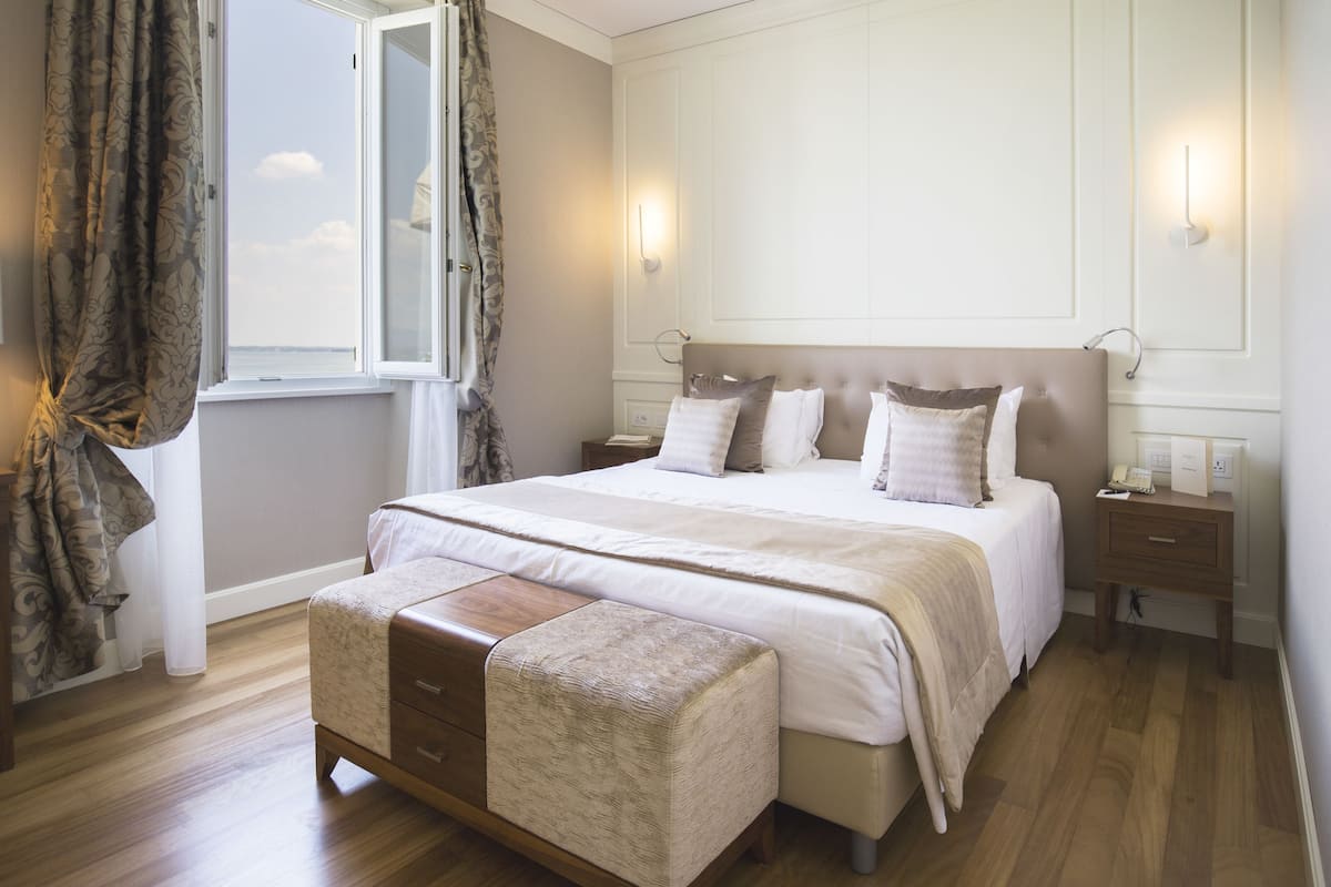Een slaapkamer met een groot bed, nachtkastjes, een houten bankje en een raam met gordijnen.