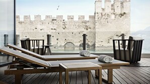 Ein Balkon mit Holzliegen und einem Tisch mit Blick auf eine historische Steinmauer und das Meer.