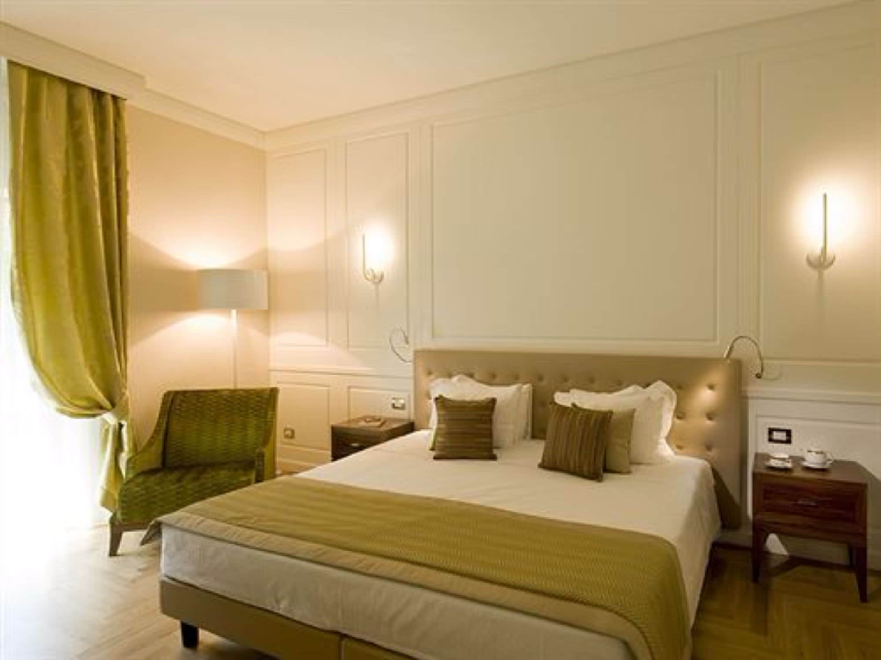 Una habitación de hotel con una cama grande, un sillón verde, una mesita de noche con una lámpara y una mesita con un reloj.