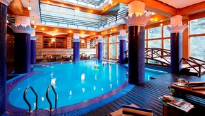 A large indoor swimming pool with a wooden deck and seating area, surrounded by tall columns and large windows.