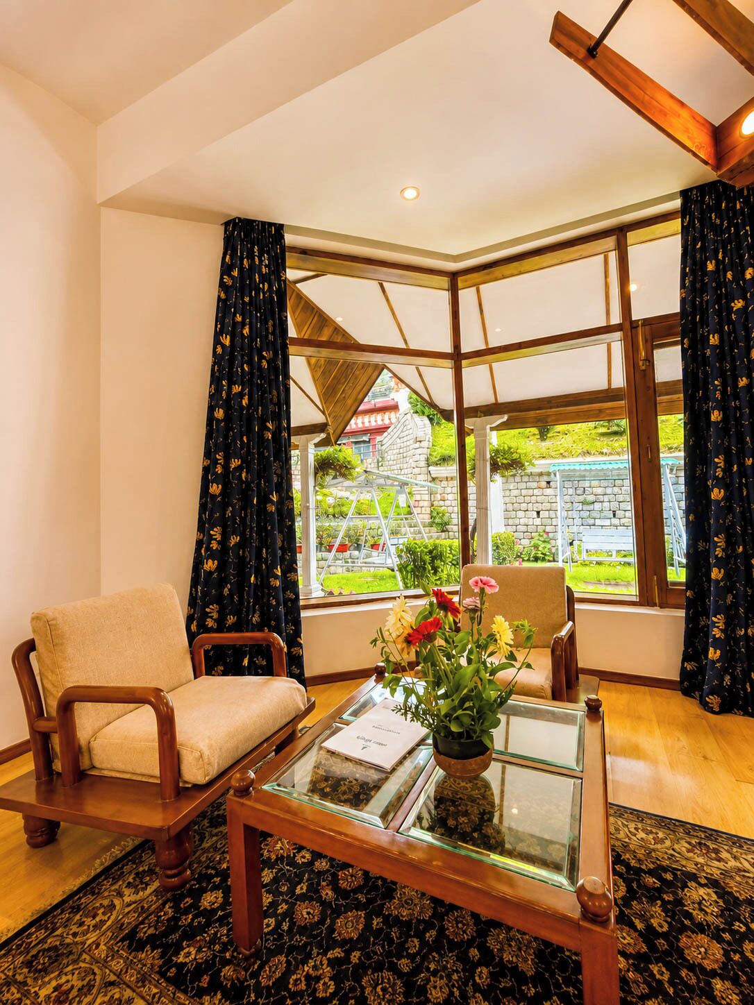 A room with a wooden chair, a glass coffee table with a vase of flowers, and a view of a garden through large windows.