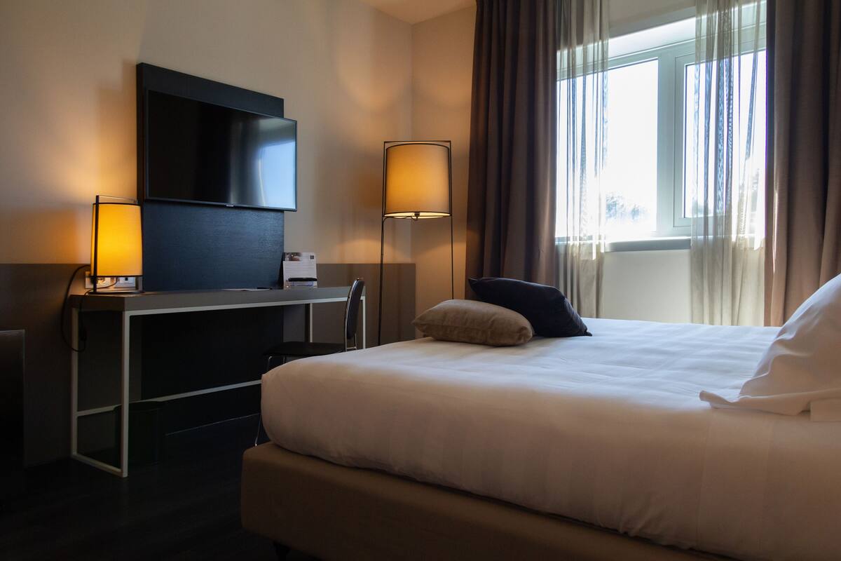A hotel room with a bed, a desk with a flat-screen TV, two lamps, and a window with curtains.