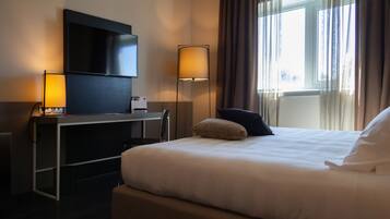 Ett hotellrum med en säng, ett skrivbord med en platt-TV, två lampor och ett fönster med gardiner.