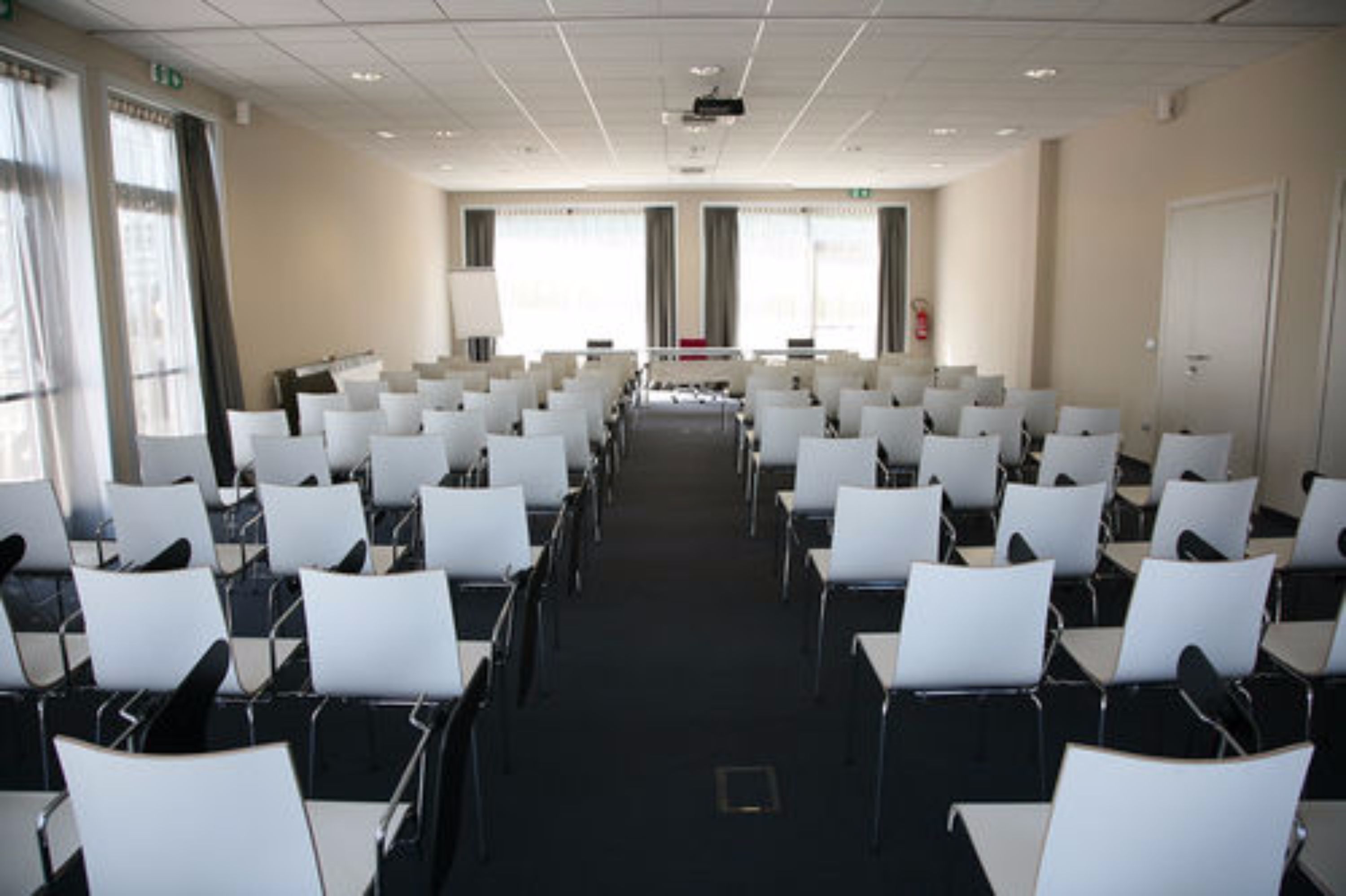 A conference room with rows of white chairs, a projector screen, and large windows.