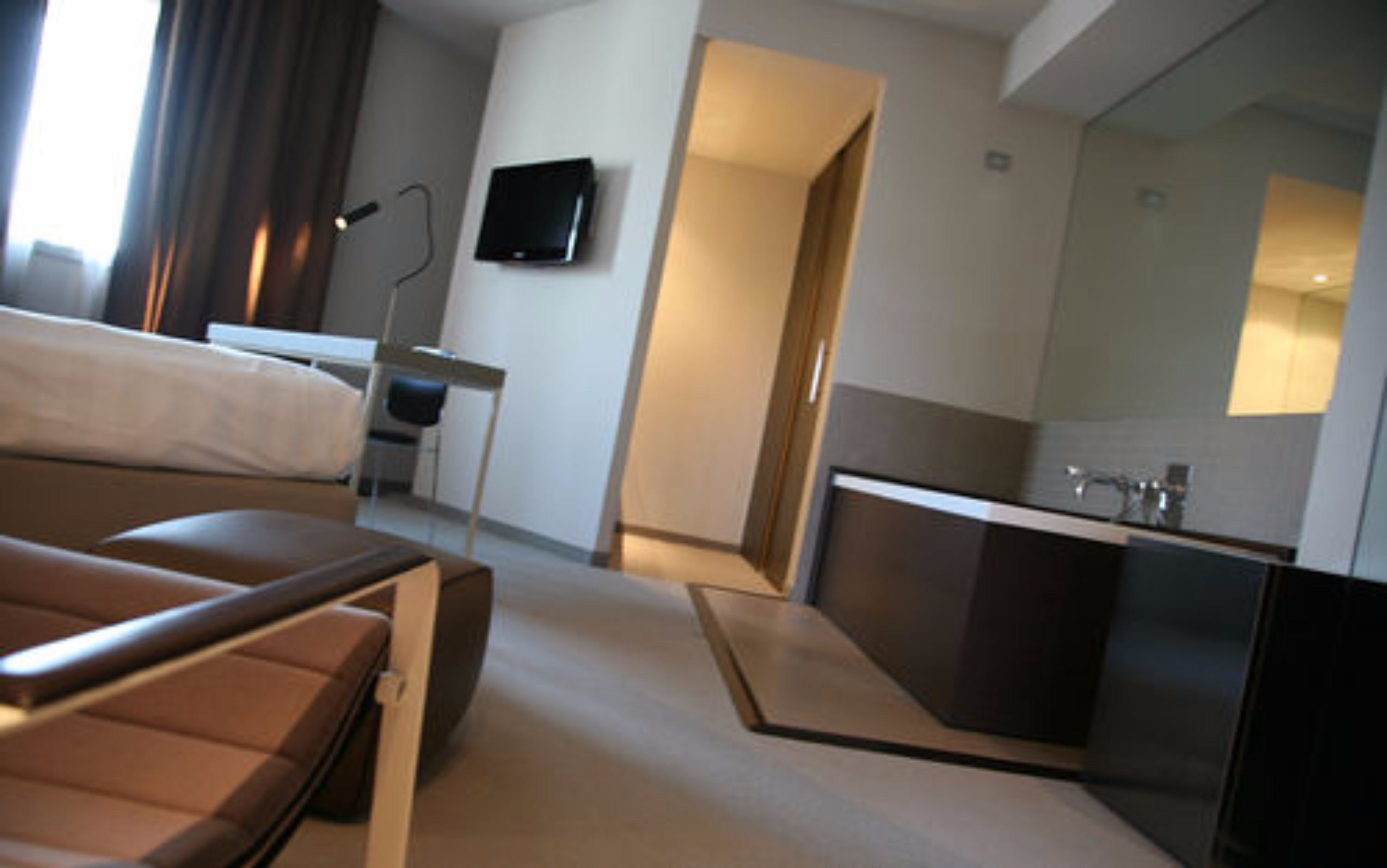 A modern hotel room with a bed, a desk, a flat-screen TV, and a kitchenette.