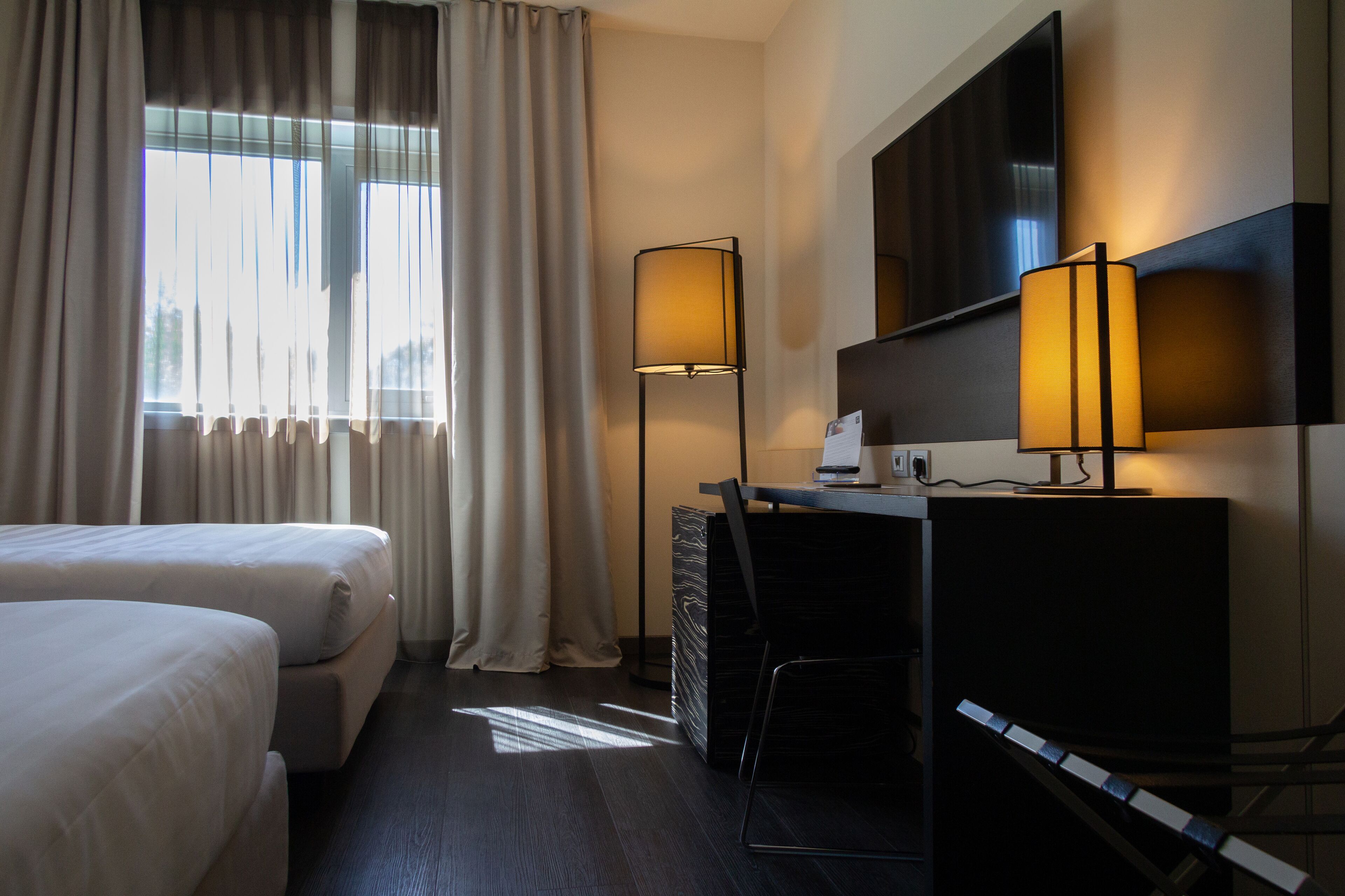 A hotel room with two beds, a desk, a chair, a flat-screen TV, and a standing lamp.