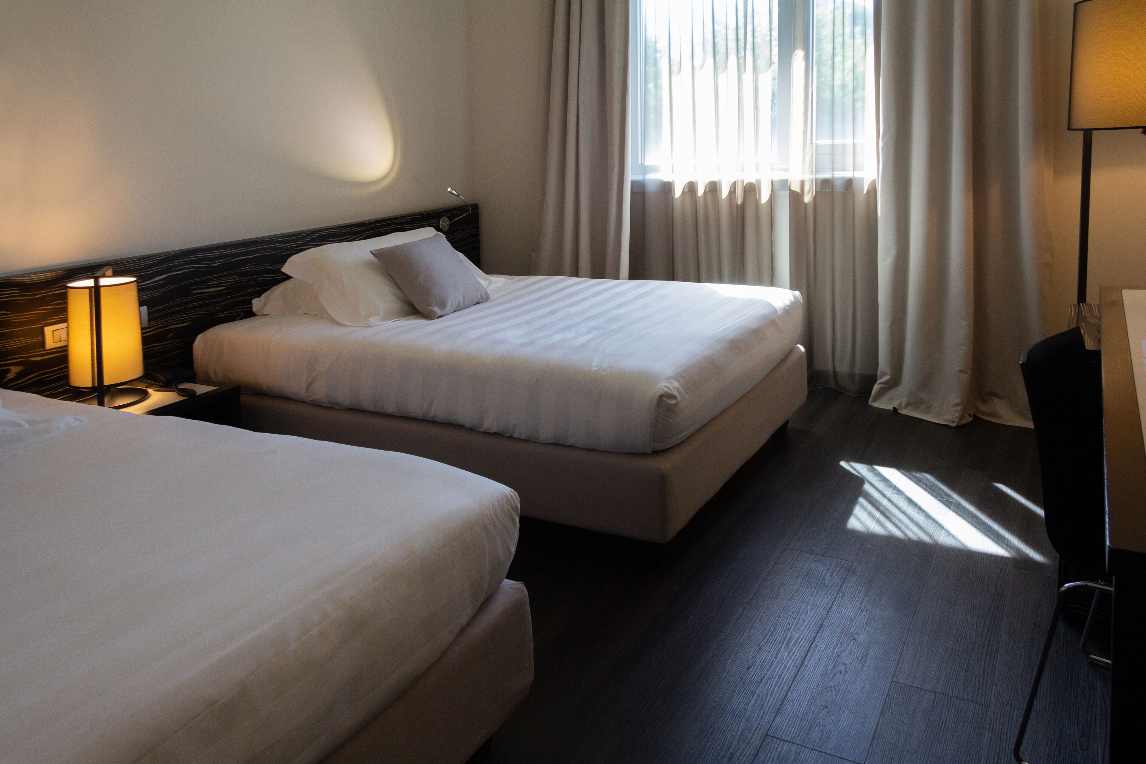 Habitación de hotel con dos camas, una mesita de noche con lámpara, un escritorio con silla y una ventana con cortinas.